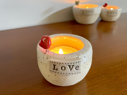 White handmade ceramic candle holder featuring tiny red cardinal and frozen pond glaze. Ceramic candle holder with 'Love' text and a lit candle.