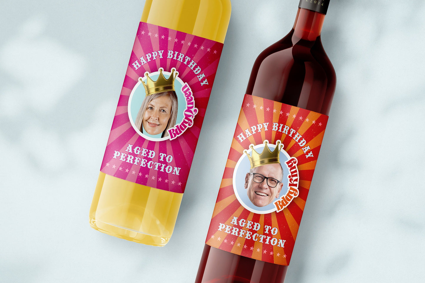 Two wine bottles with personalized labels. The labels are red and pink with a starburst design. The labels have a crown and a photo of a person. The text on the labels reads 'Happy Birthday', 'Aged to Perfection', and 'Guzzle Buddy'. 