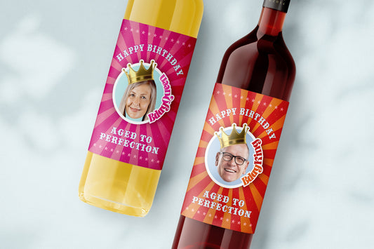 Two wine bottles with personalized labels. The labels are red and pink with a starburst design. The labels have a crown and a photo of a person. The text on the labels reads 'Happy Birthday', 'Aged to Perfection', and 'Guzzle Buddy'. 