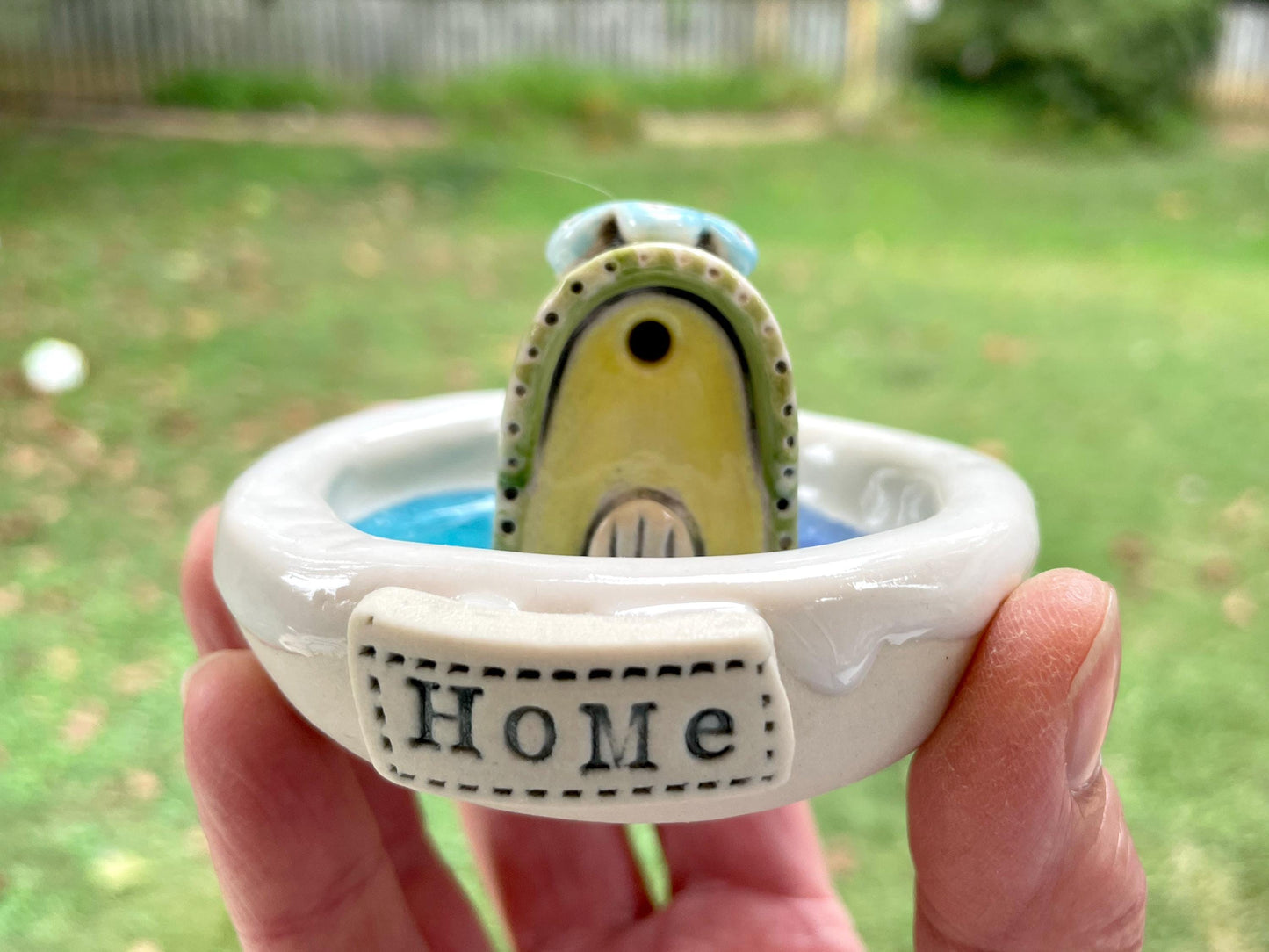 Ceramic ring with a small bird figurine and 'HOME' inscription, held in a hand outdoors.