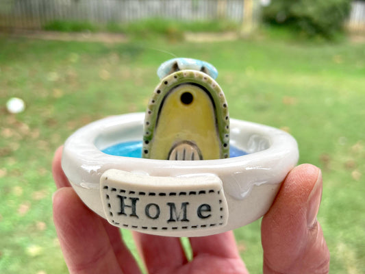 Ceramic ring with a small bird figurine and 'HOME' inscription, held in a hand outdoors.