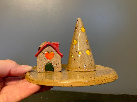 Ceramic house and cone with yellow spots on a round base held by a hand against a gray background