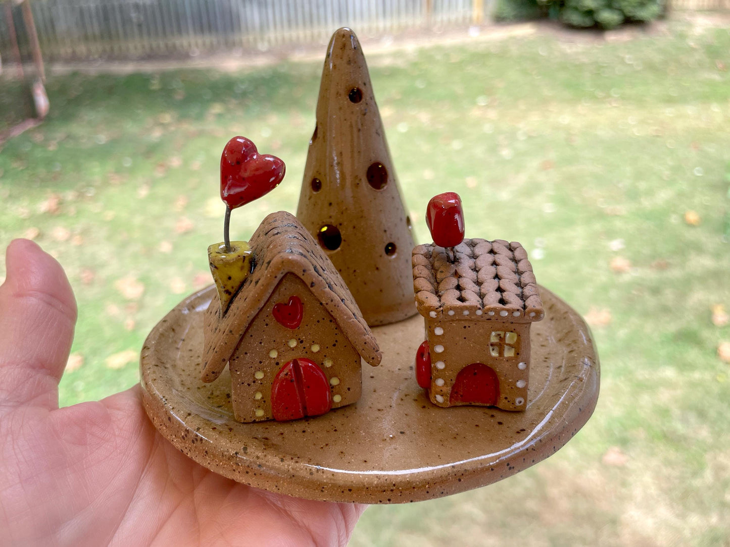 Hand holding a small ceramic plate with miniature houses and a cone-shaped object against a blurred outdoor background.