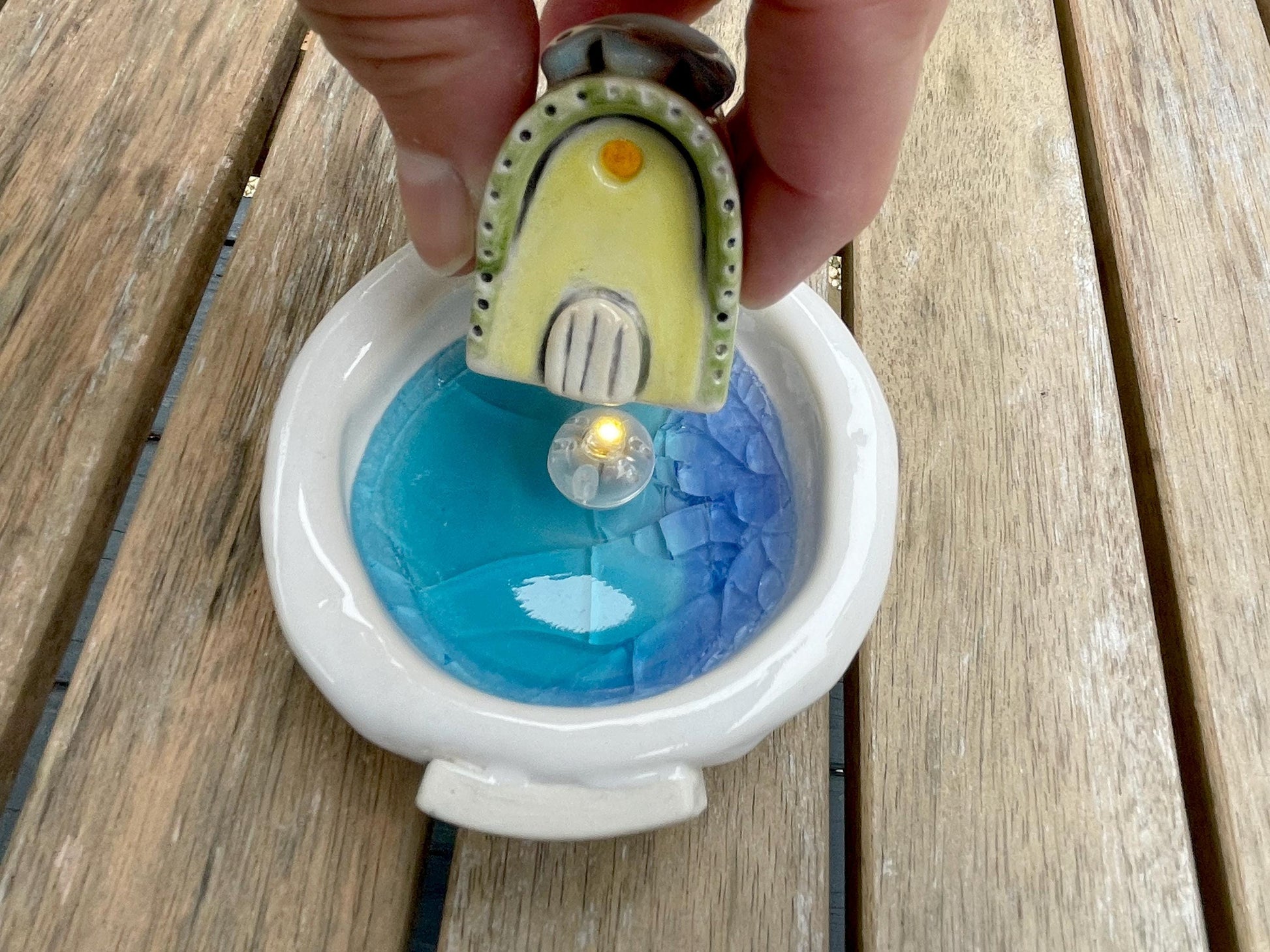 Small decorative light with a face on top, placed inside a white container with blue liquid, held by a hand on a wooden surface.