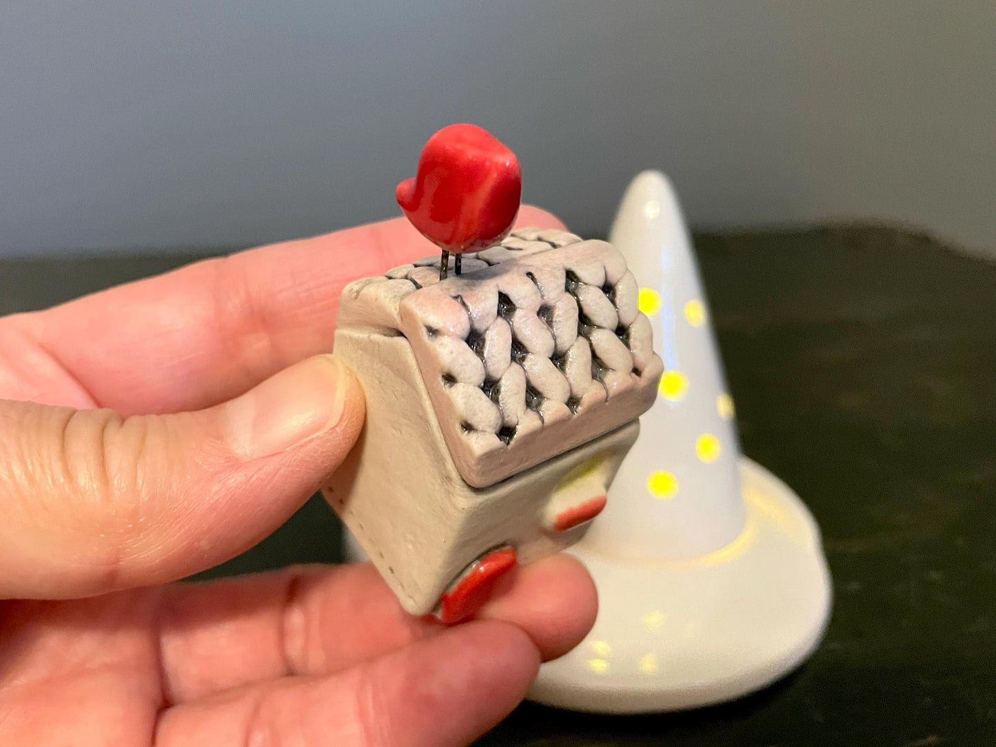 Small ceramic sculpture of a house with a red roof held by a hand, with a larger similar sculpture in the background.