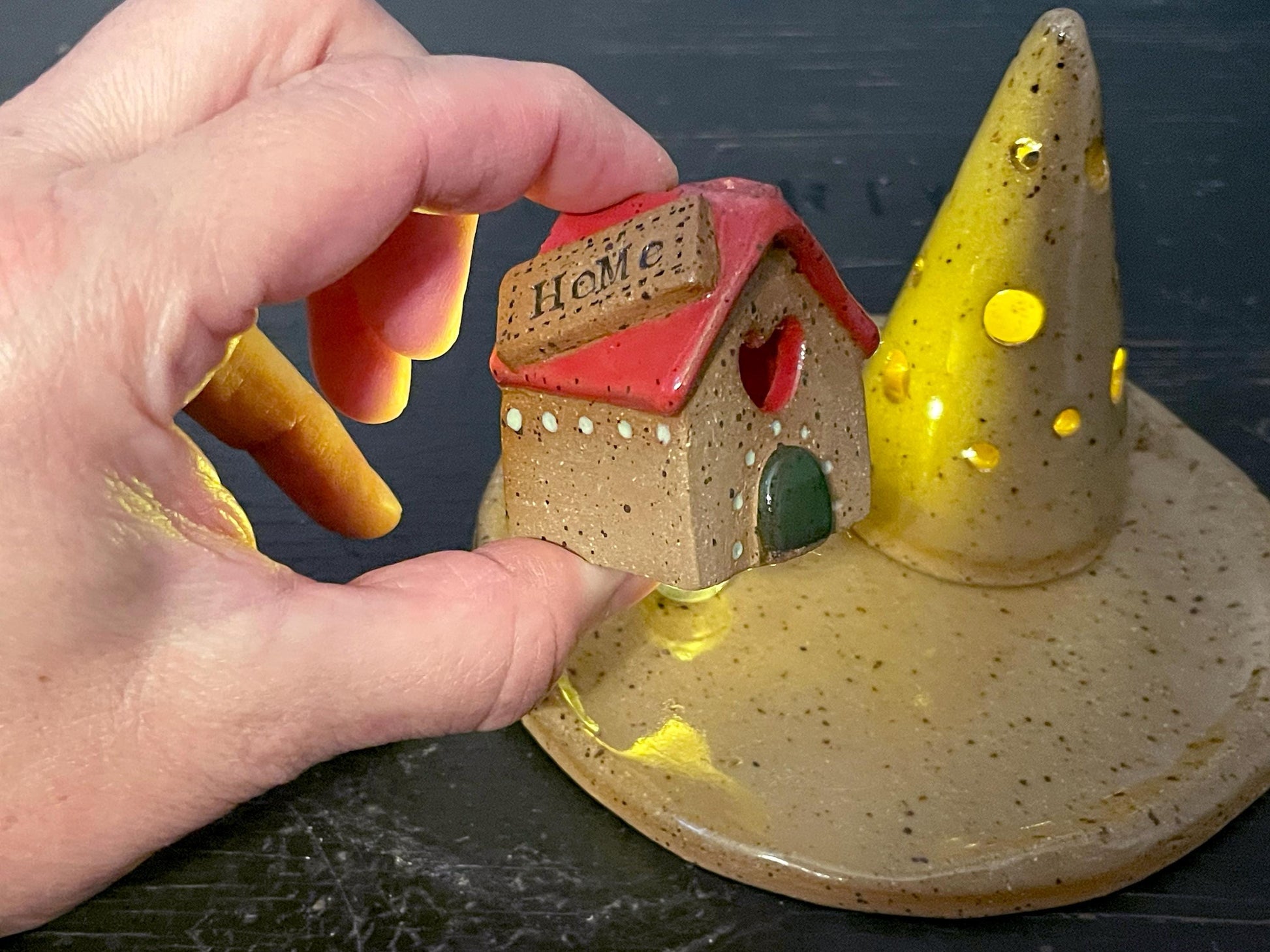 Hand holding a small ceramic house with a red roof and 'Home' text, on a dark surface.